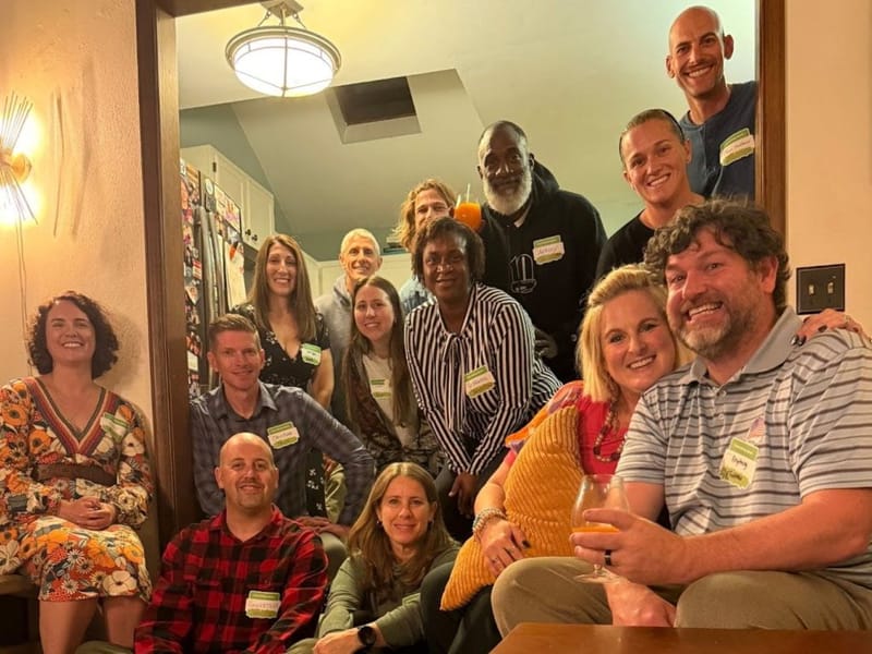 Group of about thirteen adults wearing name tags gathered closely together in a warmly lit home hallway, smiling for a group photo at a cocktail party